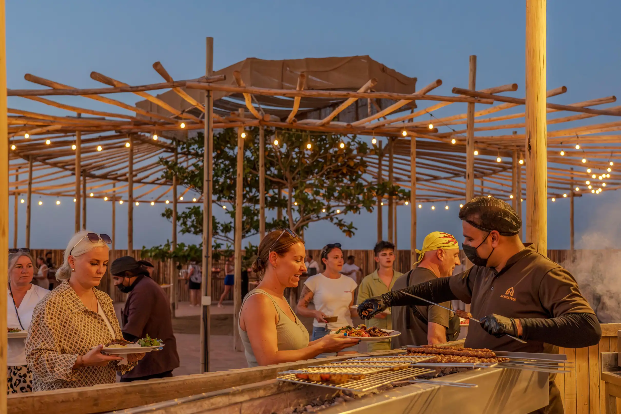 People receiving grilled food from a chef under string lights at desert safari