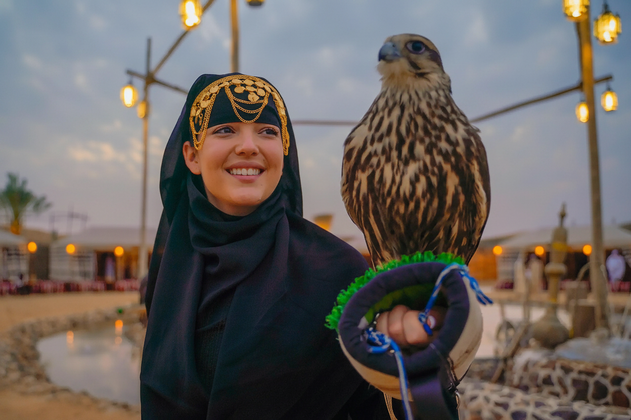 Smiling woman in traditional attire holding a falcon at a desert camp